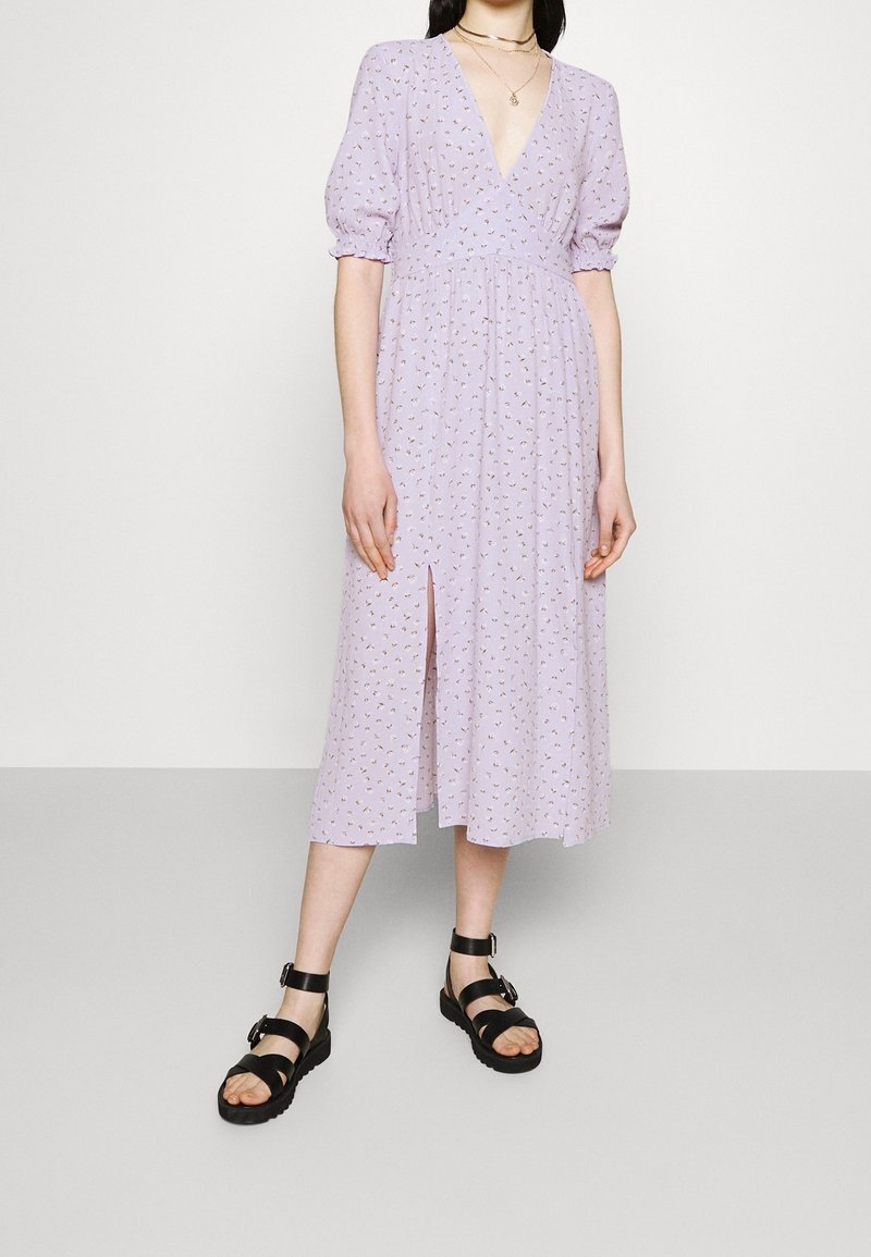 Robe midi lilas à manches bouffantes et décolleté en V profond, ornée d'un motif floral et d'une fente sur le devant. Associée à des sandales noires à lanières.