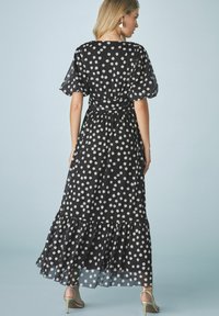 Rochie maxi neagră cu buline, cu mâneci scurte tip puff, talie legată, tiv ondulat și textură transparentă. Este decorată cu buline albe mari pe întreaga suprafață.