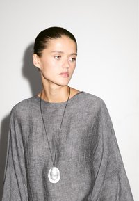 Top grigio in lino oversize con scollatura ampia, caratterizzato da una sottile texture. Un pendente ovale argentato pende da una collana con cordino nero.