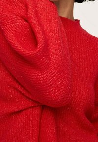 Pull en laine rouge tricoté avec une texture côtelée et une coupe ample. Le fil présente des accents de fil métallique, ajoutant une légère brillance.