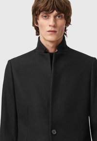 Musta villakangastakki, jossa on korkea kaulus, yksittäinen nappisulkeminen ja sileä pinta. Muotoiltu malli, jossa on rakenteelliset hartiat ja ilman taskuja.