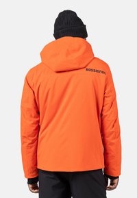 Orange vattentät jacka med huva, prydd med en svart logotyp på baksidan, designad med en smidig passform och mjuk textur. Bärs med en svart mössa.