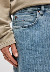 Ανοιχτό μπλε τζιν παντελόνι με διακριτικές κάθετες ρίγες, που διαθέτει μπροστινή τσέπη, χαλκομανία ριβέτα και μια μικρή κόκκινη διακοσμητική ραφή.