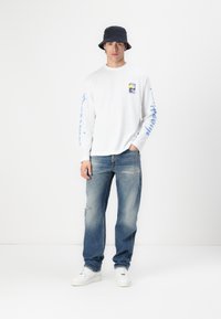 Cămașă albă cu mâneci lungi, având un grafic pe piept și un design albastru pe mâneci, asortată cu blugi deschiși la culoare, distruși, și adidași albi.