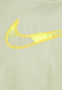 Světle zelená látka se žlutě vyšitým logem Nike ve tvaru swoosh, které obsahuje průhlednou část. Hladký povrch se jemným leskem.