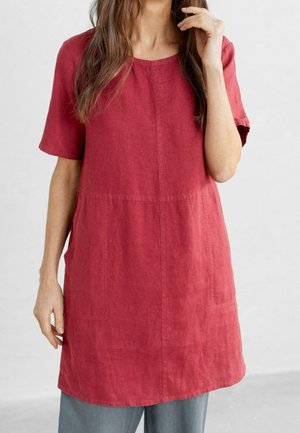 Femme portant une robe rouge ample à manches courtes et longueur genou par-dessus un pantalon gris, avec de longs cheveux bruns partiellement visibles.