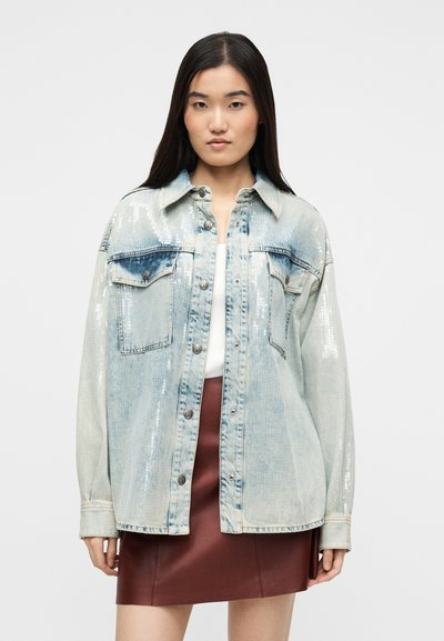 Woman wearing a light blue sequined denim jacket over a white top and a short brown leather skirt, standing against a plain background.