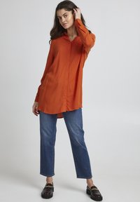 Rostfarbendes Hemd mit Knopfleiste, Kragen, langen Ärmeln und abgerundetem Saum, kombiniert mit geraden blauen Jeans und schwarzen Slippern.
