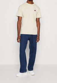 T-shirt in cotone beige con un piccolo logo sul lato sinistro del petto, abbinata a jeans in denim blu scuro e sneakers bianche. Look semplice e vestibilità rilassata.