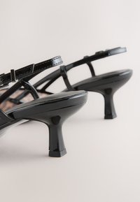 Fekete lakkbőr magas sarkú szandálok keskeny pántokkal, blokk sarokkal és texturált talppal. A kialakítás minimalista sziluettet mutat.