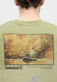 Τρία άτομα ψαρεύουν από μια μικρή βάρκα σε ήρεμο ποταμό που περιβάλλεται από φθινοπωρινά δέντρα, με το λογότυπο της Timberland και ένα απόσπασμα από κάτω.
