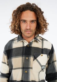 Man met krullend haar die een beige en zwart geruit button-up shirt met een borstzak draagt waarop 'Protest' staat.