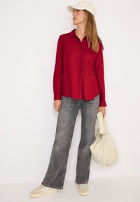 Rød knapop-shirt med lange ærmer, grå flagrende jeans, beige sneakers og en hvid kasket; modellen holder en lys taske med blød tekstur.