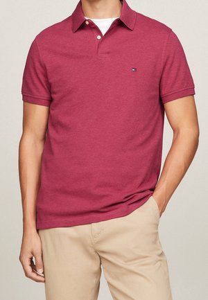 Kurzärmeliges Poloshirt aus burgunderroter Baumwolle mit klassischem Kragen, Zwei-Knopf-Leiste und kleinem Logo auf der Brust. Kombiniert mit hellbeigen Hosen.