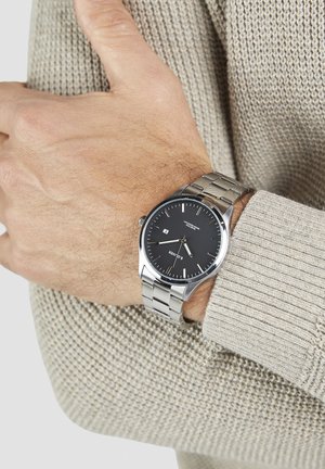 Silberne Metall-Armbanduhr mit schwarzem Zifferblatt, silbernen Stundenmarkierungen und Datumsanzeige. Getragen an einem Handgelenk vor einem strukturierten beigen Pullover.