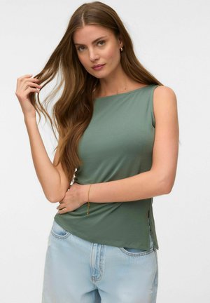 Kvinde med langt brunt hår iført ærmeløs grøn top og lyseblå jeans, poserer med hånden, der rører ved håret, mod en ensfarvet baggrund.