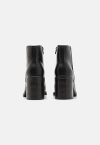 Botas de tobillo de cuero negro con punta cuadrada, un cierre vertical en la parte trasera y un tacón bloque texturizado. Acabado suave, diseño estilizado.