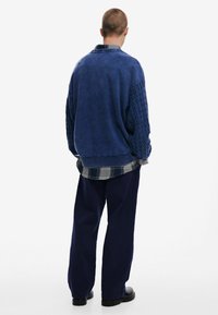 Blue textured sweater with cable-knit sleeves, layered over a plaid shirt. Paired with loose navy pants and black footwear.