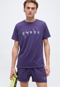 Νέος άνδρας με μωβ αθλητικά ρούχα κρατώντας ένα κίτρινο παγούρι νερού, με τα λογότυπα "HYROX" και Puma στο πουκάμισο και το σορτς.