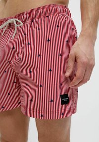 Bañador a rayas rojas con pequeños patrones de veleros en azul marino, cintura elástica con cordón y una etiqueta de la marca en negro en el lateral.