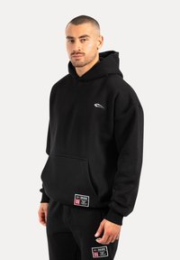 Sweat-shirt à capuche noir fabriqué dans un matériau doux, avec une poche avant, un logo sur la poitrine et des étiquettes de marque à l'ourlet.