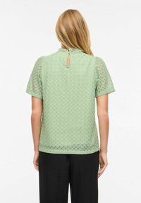 Blouse vert clair texturée avec un motif festonné, manches courtes et un détail découpé dans le dos, associée à un pantalon noir.