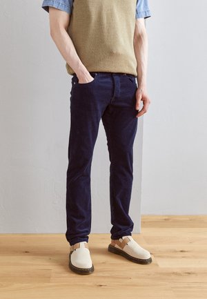 Personne portant un pull sans manches beige, une chemise bleue à manches courtes, un jean bleu foncé et des chaussures crème à enfiler, debout sur un sol en bois.
