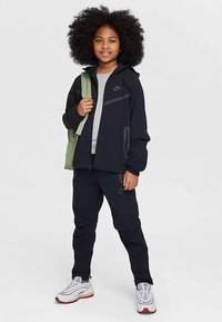 Czarna sportowa kurtka na zamek z szarymi akcentami, szara koszulka, czarne spodnie oraz białe buty sportowe z czerwonymi i szarymi akcentami. Zielony plecak zarzucony na ramię.