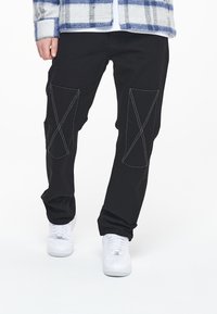 Sorte bomullsbukser med kontrasterende hvite sømmer i et 'X'-mønster, kombinert med hvite sneakers. Enkle, rette ben.