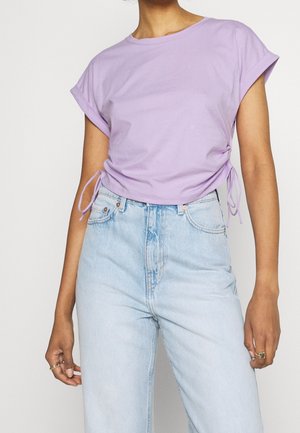 Osoba ubrana w liliową bluzkę z krótkim rękawem i wiązaniami po bokach oraz jasnoniebieskie, wysokoprofilowe dżinsy, stojąca na jednolitym tle.