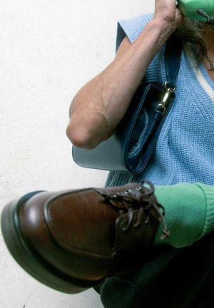 Scarpa in pelle marrone con lacci scuri, indossata con calze di un verde vivace. Borsa blu tenuta da una mano, con una maglia blu a maglia visibile.