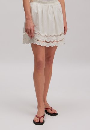 Femme portant une jupe blanche avec un ourlet en dentelle festonné et des sandales rouges à lanières, debout devant un fond gris uni.