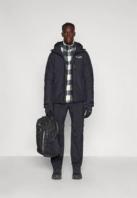 Man gekleed in een zwarte winterjack, geruite shirt, zwarte broek, handschoenen, met een zwarte rugzak tegen een effen achtergrond.