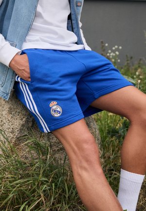 Personne assise à l’extérieur portant un short bleu du Real Madrid, des chaussettes blanches, une chemise blanche et un gilet en jean, les mains dans les poches.
