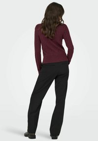 Burgundijas krāsa adīta džemperis ar ribinātu apakšmalu, kombinēts ar melnām taisnām biksēm un melnām apaviem, skatīts no aizmugures.