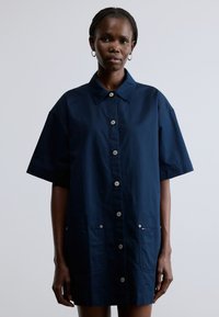 Tengerészkék, oversized szabású, rövid ujjú ingruha gombos elejjel, két elülső zsebbel és galléros nyakkal. Sima anyagtextúra.