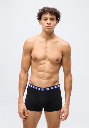 Jeune homme torse nu aux cheveux bouclés, portant un boxer noir avec une ceinture bleu marine, face à l'avant sur un fond uni.