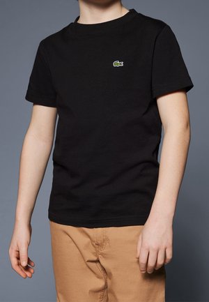 Barn iført en sort kortærmet t-shirt med en lille grøn krokodillelogo på brystet og beige bukser, stående mod en grå baggrund.