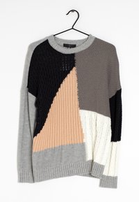 Pull tricoté avec un design à sections mélangées en gris, noir, beige et blanc. Présente des poignets côtelés et des textures variées. Suspendu à un crochet noir.