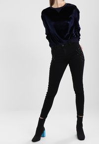Top lungo in velluto blu navy con scollatura rotonda abbinato a jeans neri a vita alta con borchie argentate. Stivaletti neri con tacchi blu.