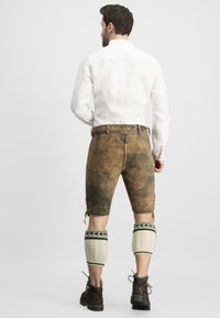 Braune Lederhosen mit dekorativen seitlichen Schnürungen, kombiniert mit knielangen, weißen gestrickten Beinstulpen mit grünen Akzenten und braunen Schuhen.