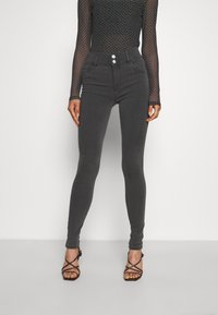 Jeans skinny de cintura alta, cinza escuro, com duplo botão e bolsos frontais, combinados com sandálias de salto com tiras. Textura de tecido suave.