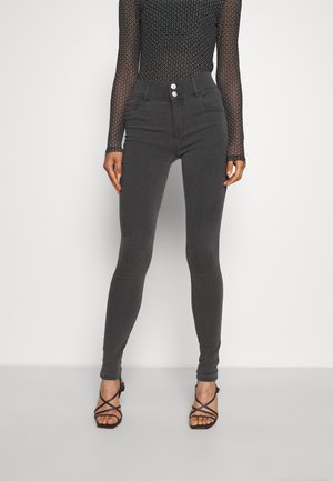 Jeans skinny de cintura alta, cinza escuro, com duplo botão e bolsos frontais, combinados com sandálias de salto com tiras. Textura de tecido suave.