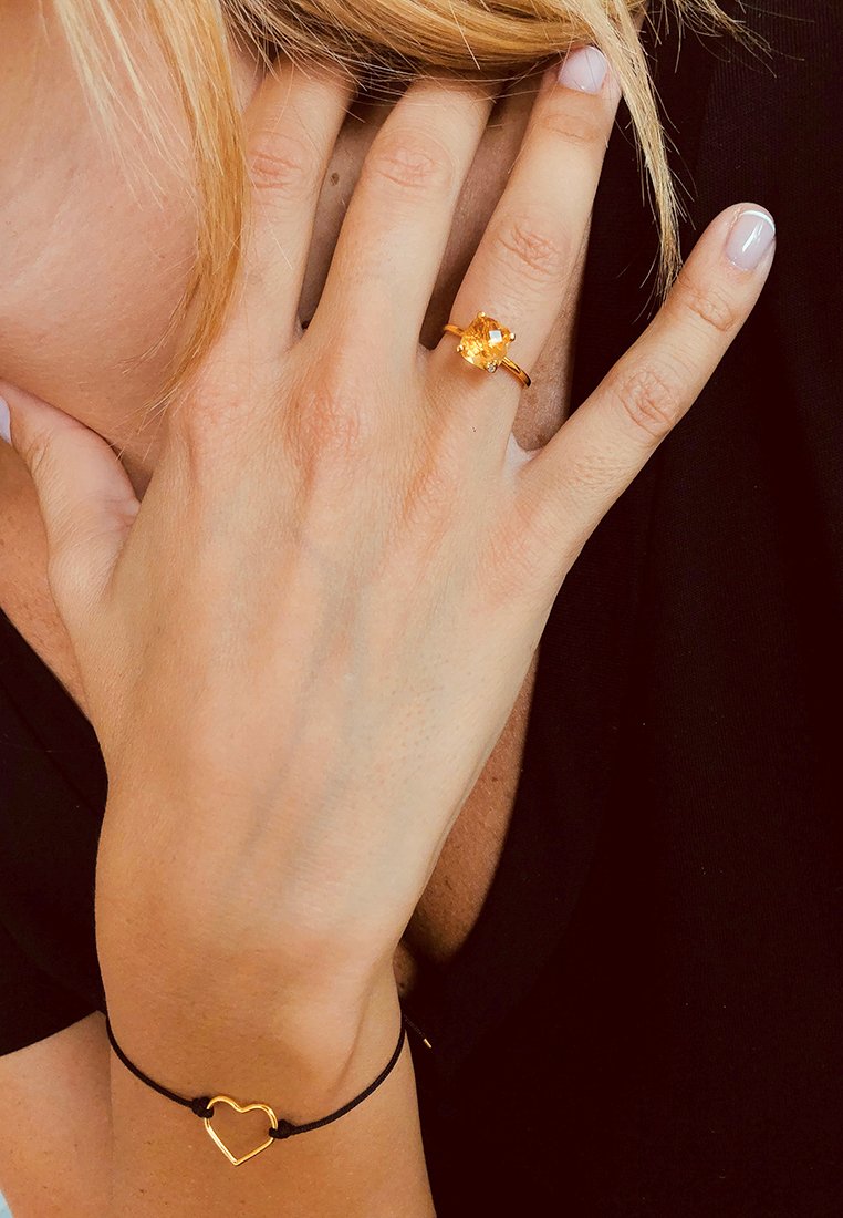 Dłoń z złotym pierścionkiem z żółtym kamieniem spoczywająca w pobliżu ucha osoby o blond włosach, noszącej czarną bransoletkę w kształcie serca i czarną bluzkę.