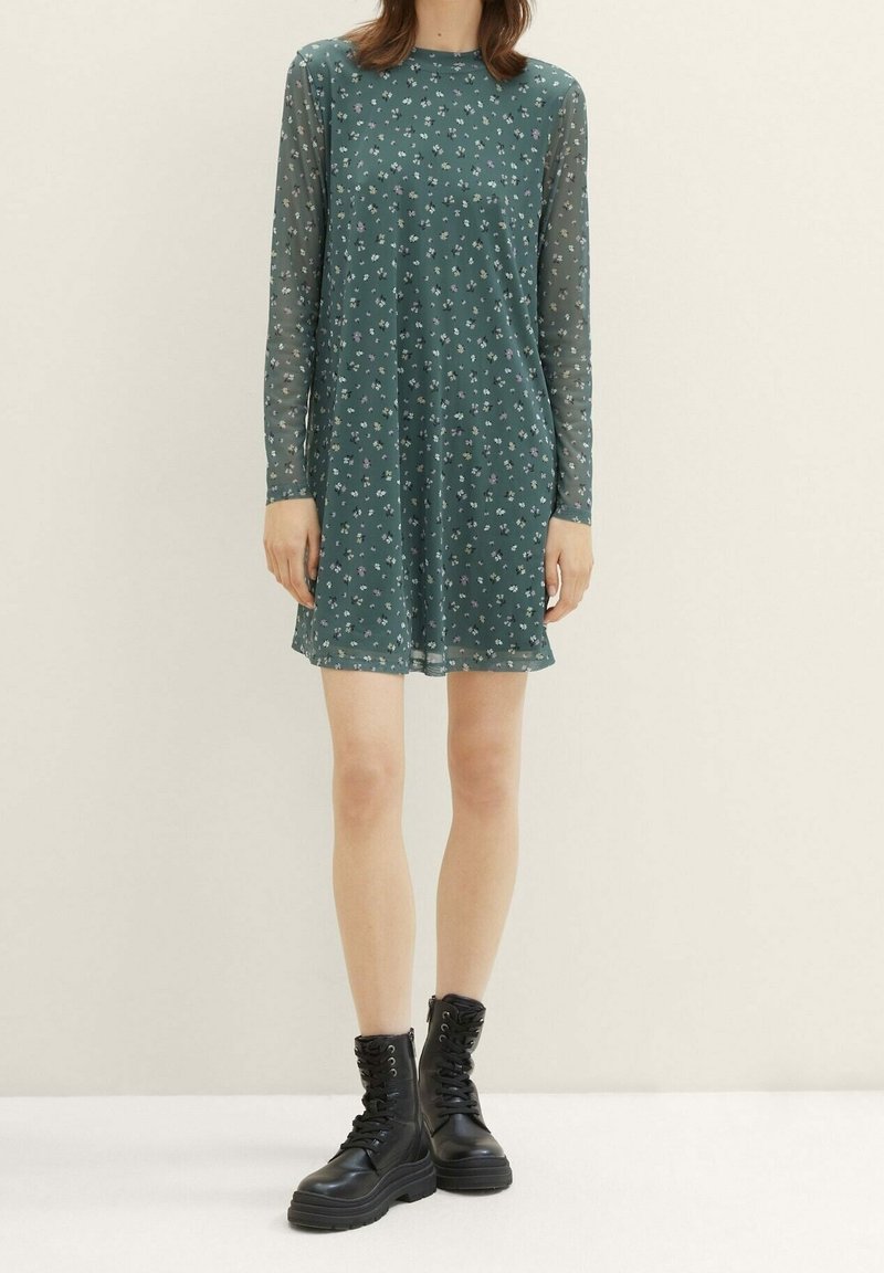 Robe florale verte avec manches longues en tissu transparent, encolure ronde, et coupe ample. Portée avec des bottes de combat noires à lacets.