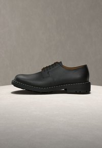 Chaussure habillée en cuir noir à lacets avec une surface texturée, une semelle épaisse et une couture blanche visible le long du bord sur un fond uni.