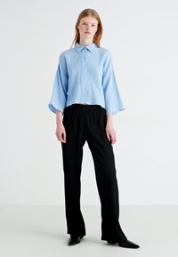 Camicetta azzurrina cortata in tessuto testurizzato con chiusura a bottone; abbinata a pantaloni neri dalla vestibilità comoda e scarpe nere eleganti.