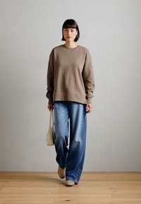 Overdimensjonert brun hettegenser kombinert med vide blåjeans. Holder en liten beige veske. Teksturert stoff, avslappet passform, subtil merking på ermet.