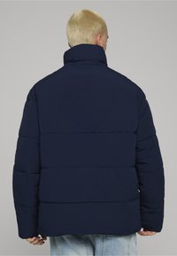 Marineblå dunjakke med høy krage og quiltet design, med et ton-i-ton logodetalj på baksiden. Myk, glatt stofftekstur.