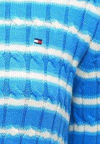 Pull tricoté à rayures bleues et blanches avec un motif en torsades, agrémenté d'un petit patch logo aux couleurs rouge, blanc et bleu.